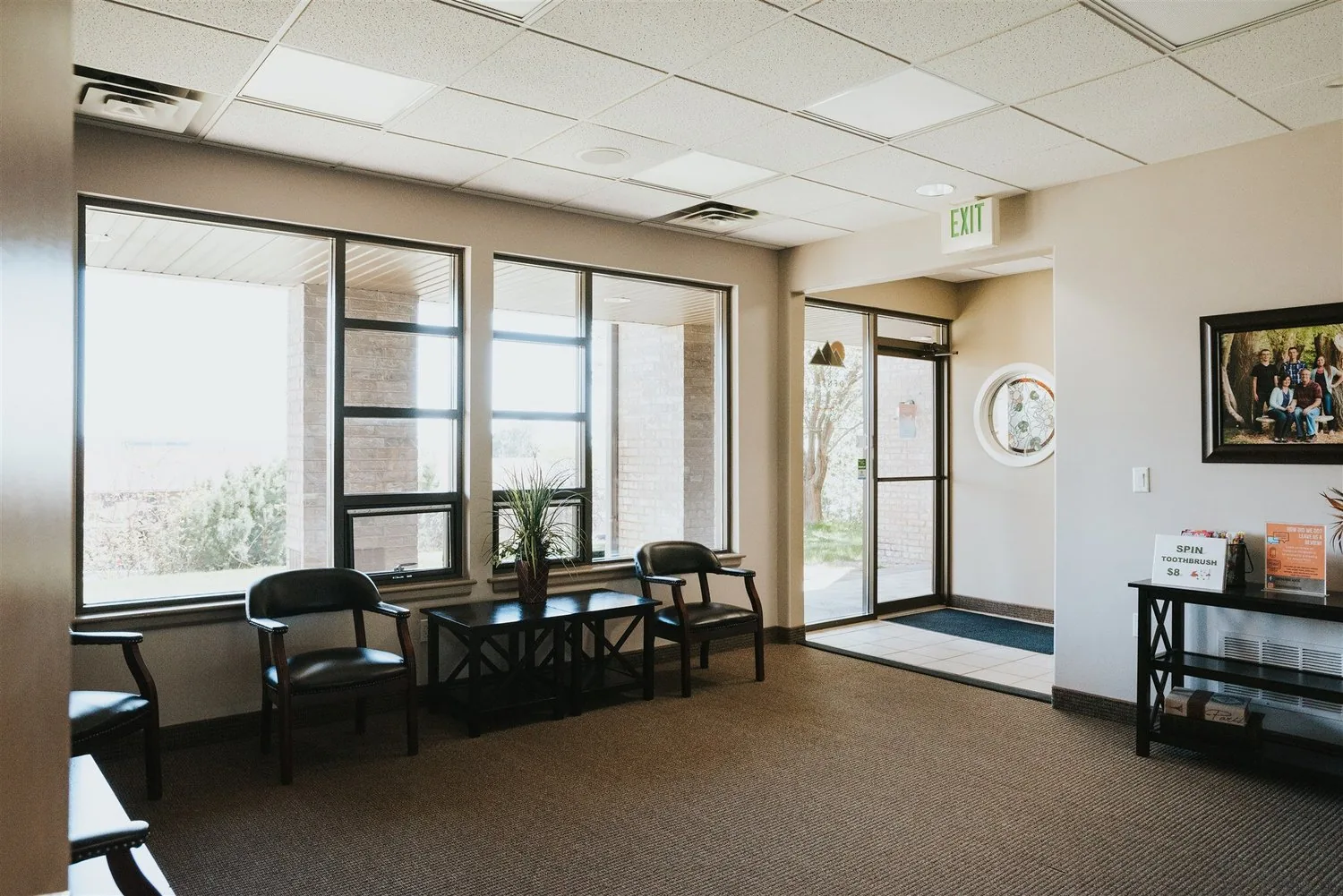 Waiting room with armchairs beside large windows, a small table with a potted plant, and glass entrance doors.